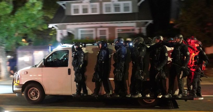 Portland police in riot gear are pictured riding on the sideboards of a police van in an Aug. 8 file photo after dispersing a mob in front of the Mutnomah County Sheriff's Office.