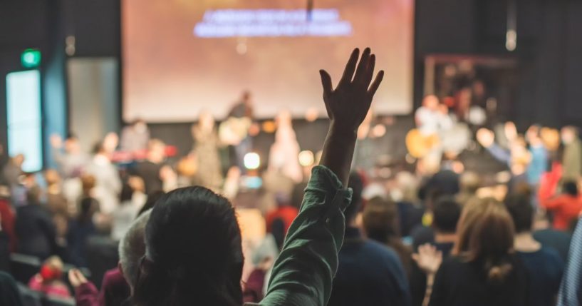 A church congregation worships through music during the service. The back of one woman is in focus, and she is raising a hand in praise.