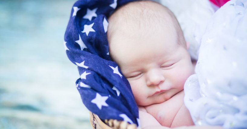 This image shows a newborn baby sleeping in a basket filled with patriotic scarves.