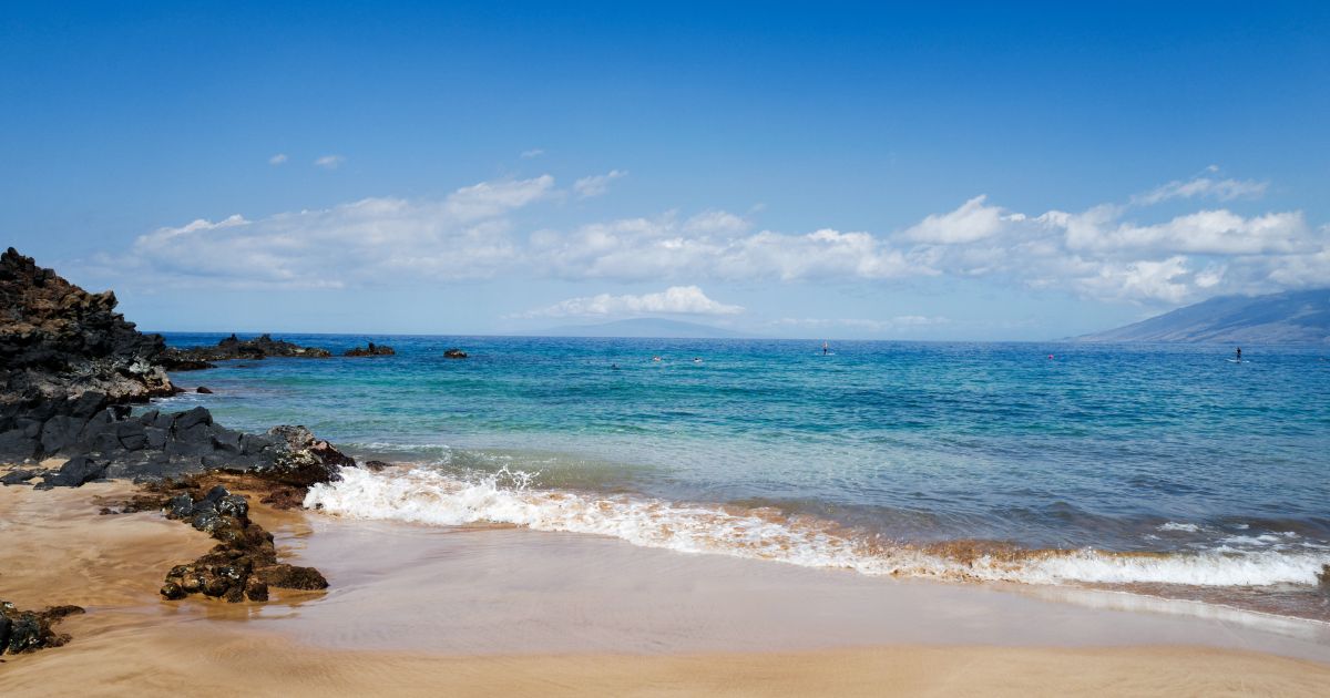 On a sunny day at Wailea beach, small waves break on the sand near volcanic rocks as tourists snorkel and use stand up paddleboards, Wailea, Maui, Hawaii, 2016.