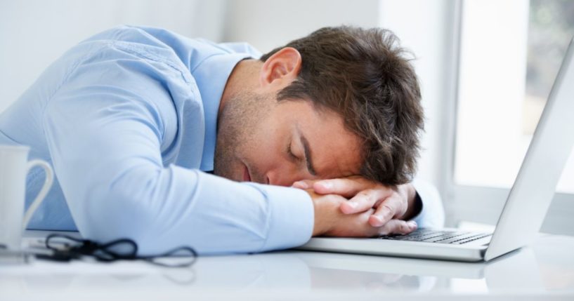 This image shows a businessman asleep on his laptop on his desk at work.